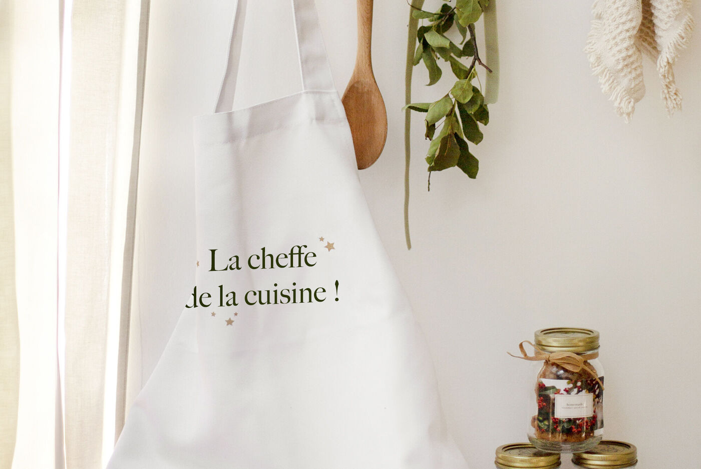 Un tablier blanc avec l'inscription "La cheffe de la cuisine!" accroché entre une cuillère en bois et des bocaux d'ingrédients.